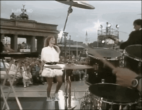 a woman in a white skirt is dancing on a stage with a man playing drums behind her