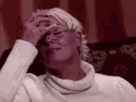 a woman wearing glasses and a white sweater is sitting on a couch covering her face with her hand .
