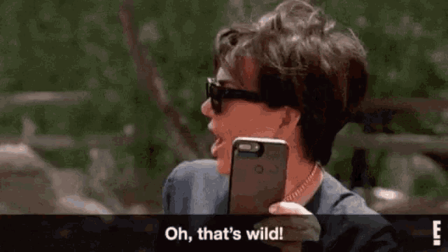 a woman wearing sunglasses is holding a cell phone and saying oh that 's wild .