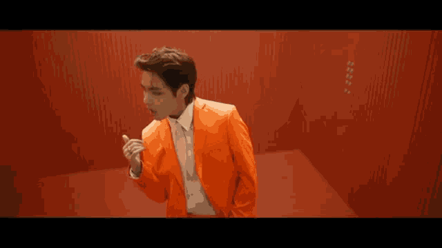 a man in an orange suit and white shirt is standing in front of a red wall .