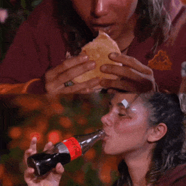 a woman drinking a bottle of coca cola next to a woman eating a sandwich