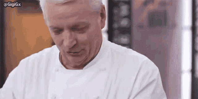a man in a white chef 's coat is looking down at something in a kitchen .