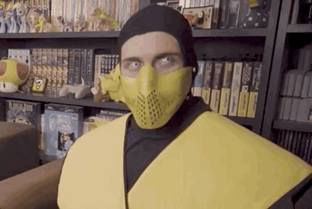 a man wearing a yellow mask and a black hat is standing in front of a bookshelf filled with books .