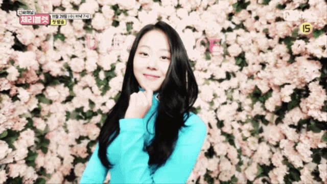 a woman in a blue shirt stands in front of a wall of pink flowers with the number 15 on the bottom