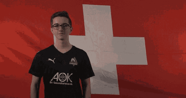 a man wearing a black aok shirt stands in front of a large swiss flag
