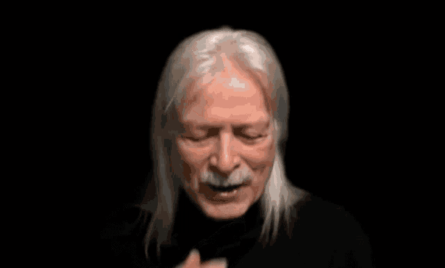 a man with long white hair and a mustache is smiling in the dark .