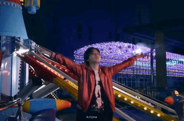 a man in a red jacket is standing in front of a roller coaster at night