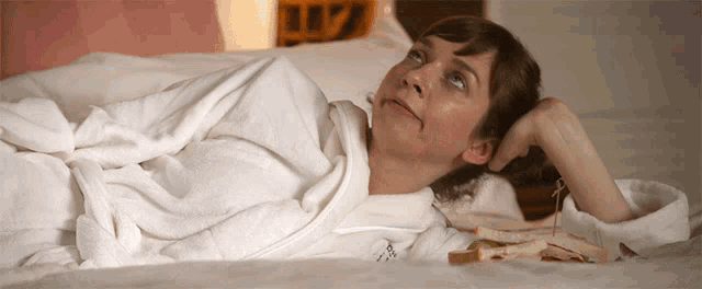 a woman in a bathrobe is laying on a bed with a sandwich on the bed