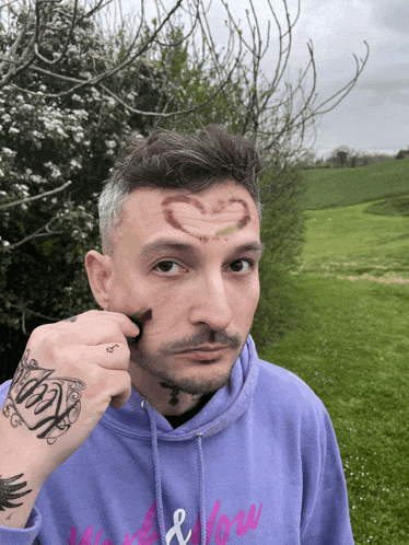 a man with a heart drawn on his forehead is wearing a purple hoodie with the word love on it