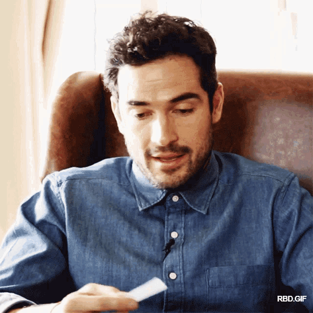 a man in a denim shirt is sitting in a chair looking at a piece of paper