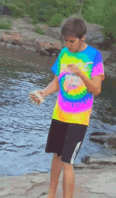 a boy in a colorful tie dye shirt stands in front of a river