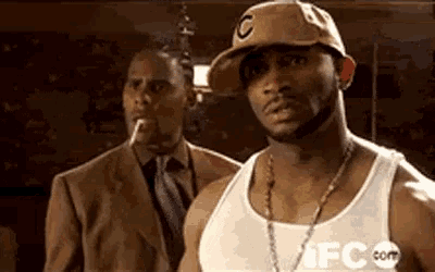 a man in a hat with the letter c on it is standing next to another man in a suit smoking a cigarette .