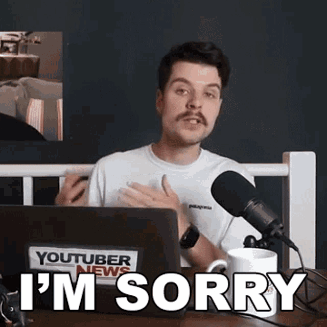 a man with a mustache is sitting in front of a microphone with the words i 'm sorry on the table .