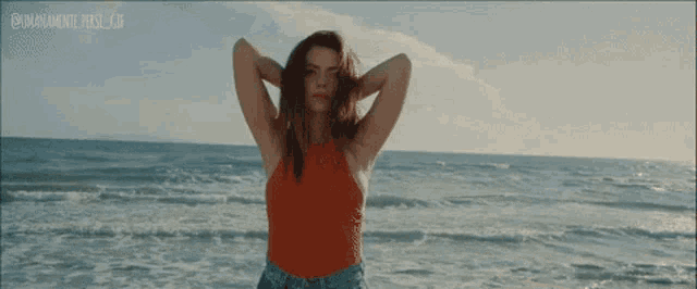 a woman in a red tank top stands on the beach with her hands behind her head