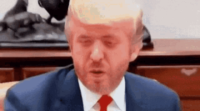 donald trump is wearing a suit and tie and making a funny face while sitting at a table .