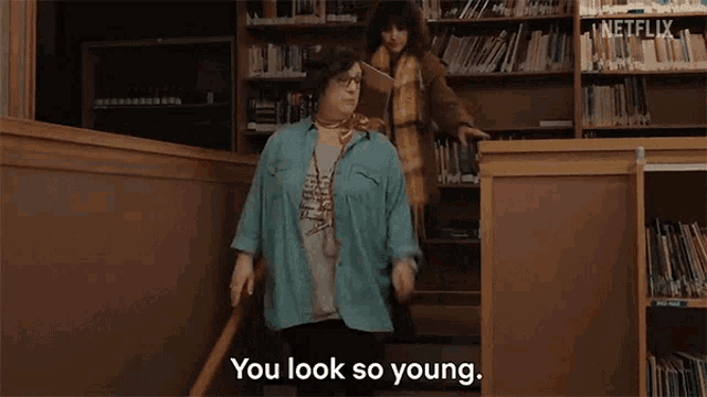 a woman is walking down stairs in a library while another woman says `` you look so young '' .