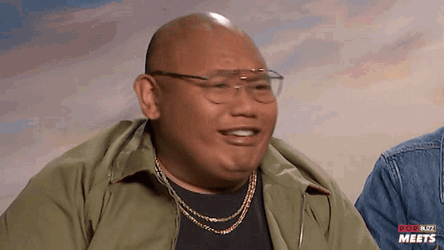 a bald man wearing glasses and a green jacket is smiling in front of a pop buzz meets sign