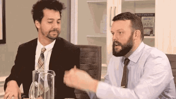 two men are sitting at a table talking to each other . one of the men is wearing a suit and tie .
