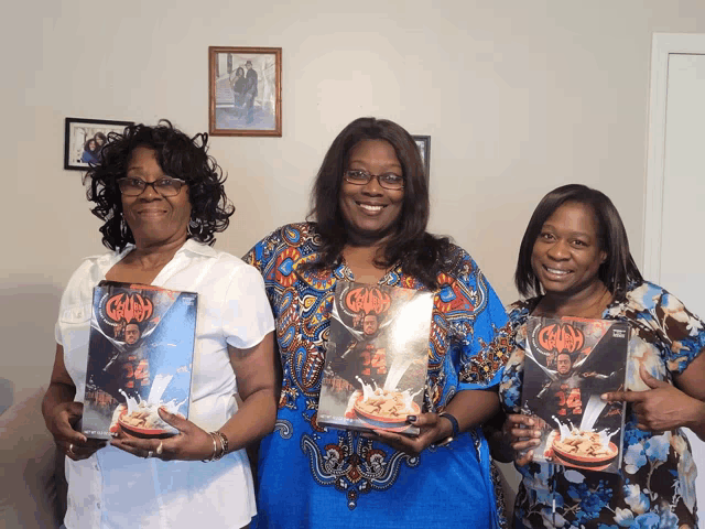 three women are posing for a picture and one of them is holding a cereal box that has the word cereal on it