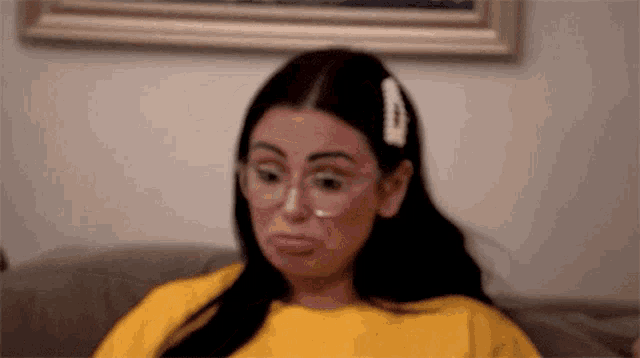 a woman wearing glasses and a yellow shirt is sitting on a couch making a face .