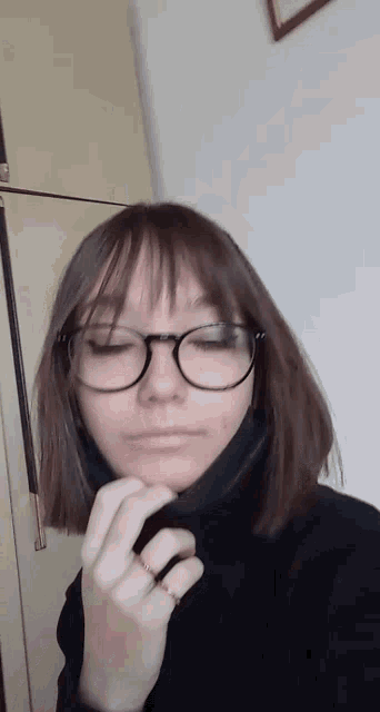 a girl wearing glasses and a black turtleneck is taking a selfie with her eyes closed .