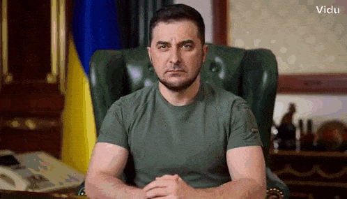 a man in a green shirt is sitting in a chair with his hands folded .
