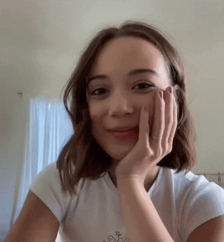 a woman in a white t-shirt is holding her hand to her face and smiling .