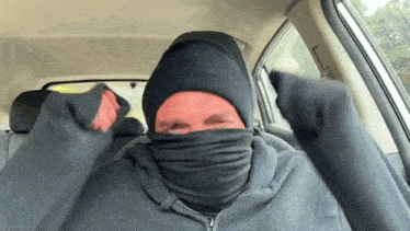 a man wearing a mask and a beanie is sitting in a car with his arms outstretched .