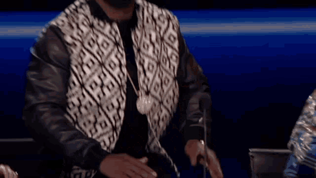 a man wearing a black and white jacket and a necklace is sitting in front of a microphone on a stage .