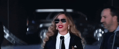 a woman wearing sunglasses , a suit and tie , and red lipstick is walking down a street .