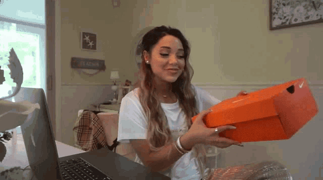 a woman sitting in front of a laptop is holding an orange nike box