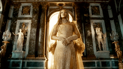 a woman in a gold dress and gloves stands in a room with statues