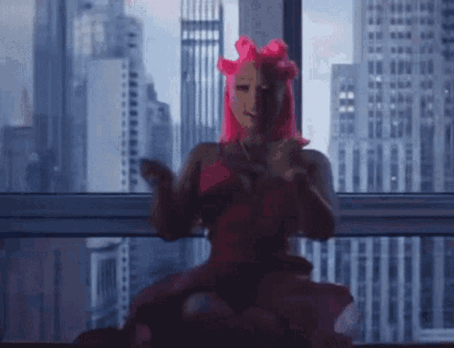 a woman with pink hair is sitting in front of a window with a city skyline in the background