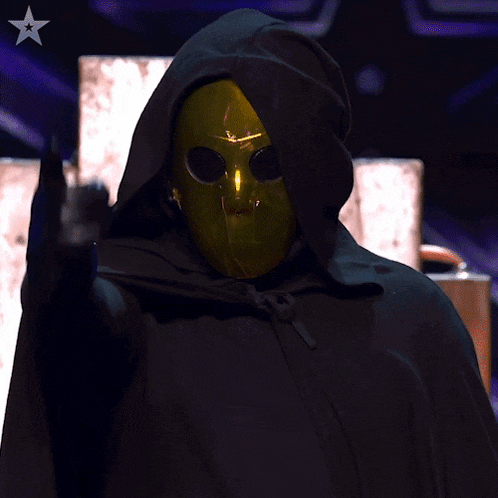 a person wearing a black hooded cape and a gold mask pointing at the camera