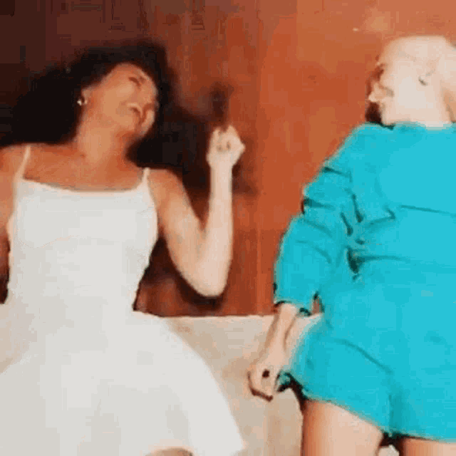 two women are dancing on a couch and one is wearing a white dress and the other is wearing blue shorts .