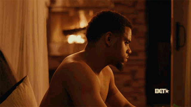 a shirtless man sits in front of a fireplace with a bet logo behind him