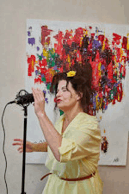 a woman in a yellow dress singing into a microphone in front of a colorful painting