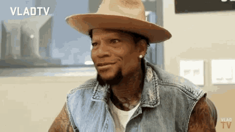 a man wearing a hat and a denim vest is looking at the camera .