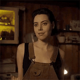 a woman wearing overalls and a black tank top is standing in a room .
