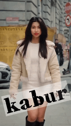 a woman wearing a white jacket and a white dress is standing in front of a sign that says " kabur " on it