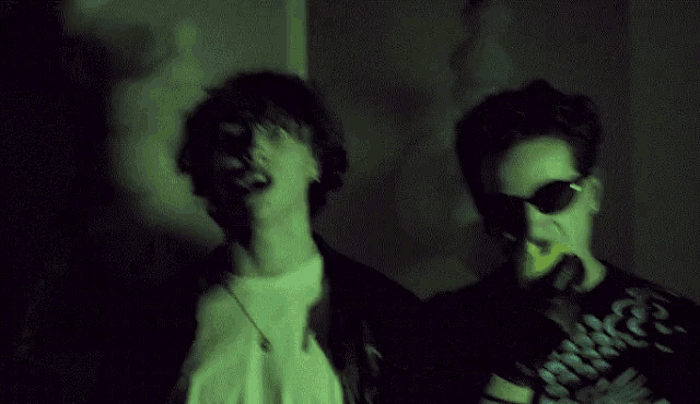 two men are standing next to each other in a dark room . one of the men is wearing sunglasses and holding an apple .