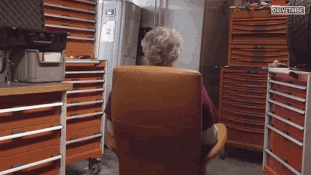 an older woman sits in a chair in a garage with a sign that says drivetribe on it