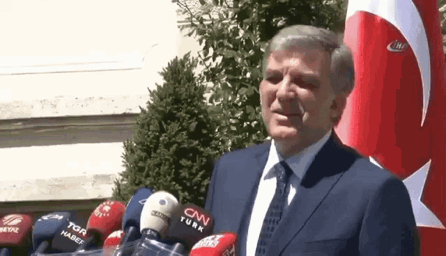 a man in a suit and tie stands in front of a row of microphones including cnn turk
