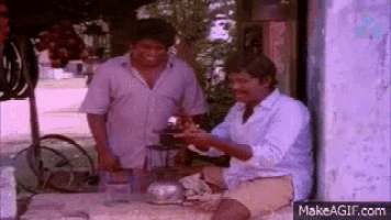two men are sitting at a table in front of a building . one of the men is holding a plate of food .