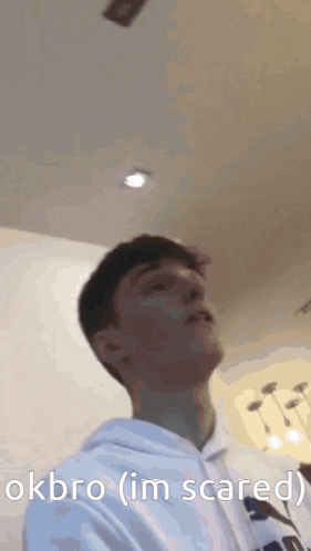 a man in a white hoodie is standing in front of a ceiling and says okbro ( i 'm scared )