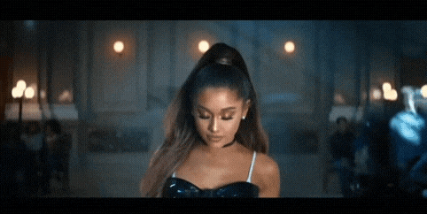 ariana grande is wearing a black dress and a ponytail while standing in a dark room .