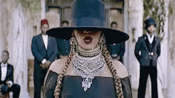 a woman wearing a hat and necklace is standing in front of a group of men in suits .