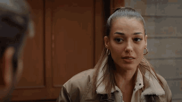 a woman in a brown jacket and hoop earrings is looking at a man in a room .
