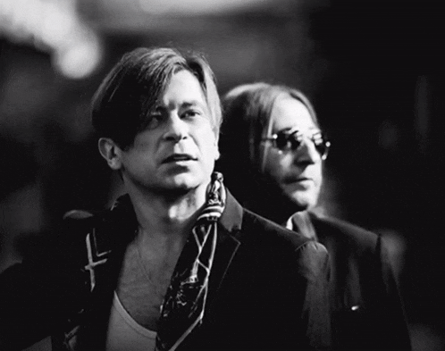a man with a scarf around his neck stands next to another man in a suit