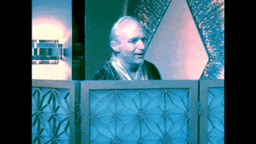 a man in a robe stands behind a blue geometric screen